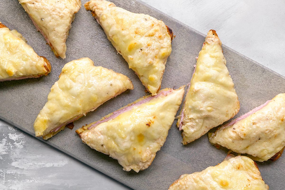 Several golden, triangular sandwiches with melted cheese on top are arranged on a gray serving tray. The sandwiches appear toasted and filled, possibly with ham and cheese.