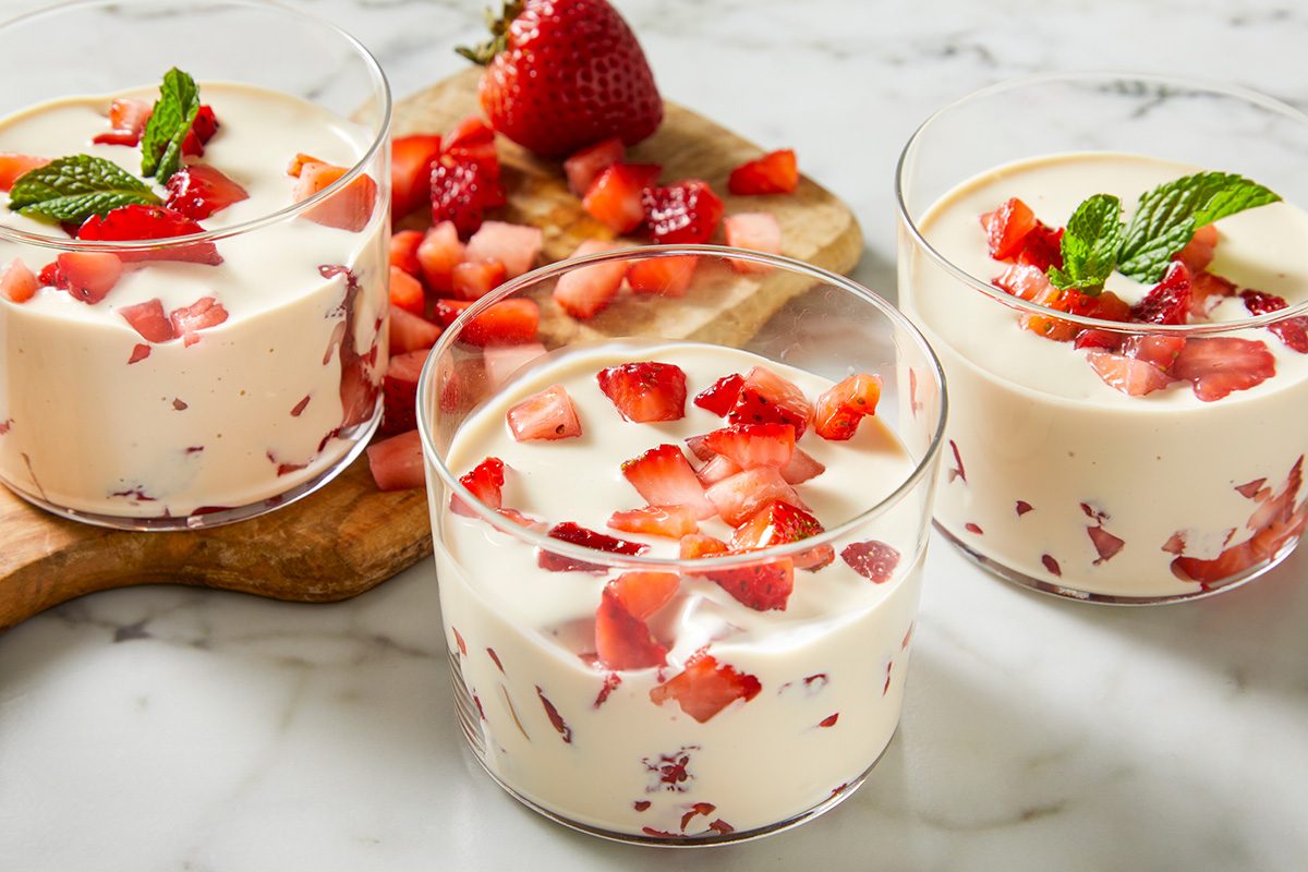 Three glass cups filled with creamy dessert mixed with chopped strawberries, topped with fresh mint leaves. Extra diced strawberries and a whole strawberry are on a wooden board in the background.