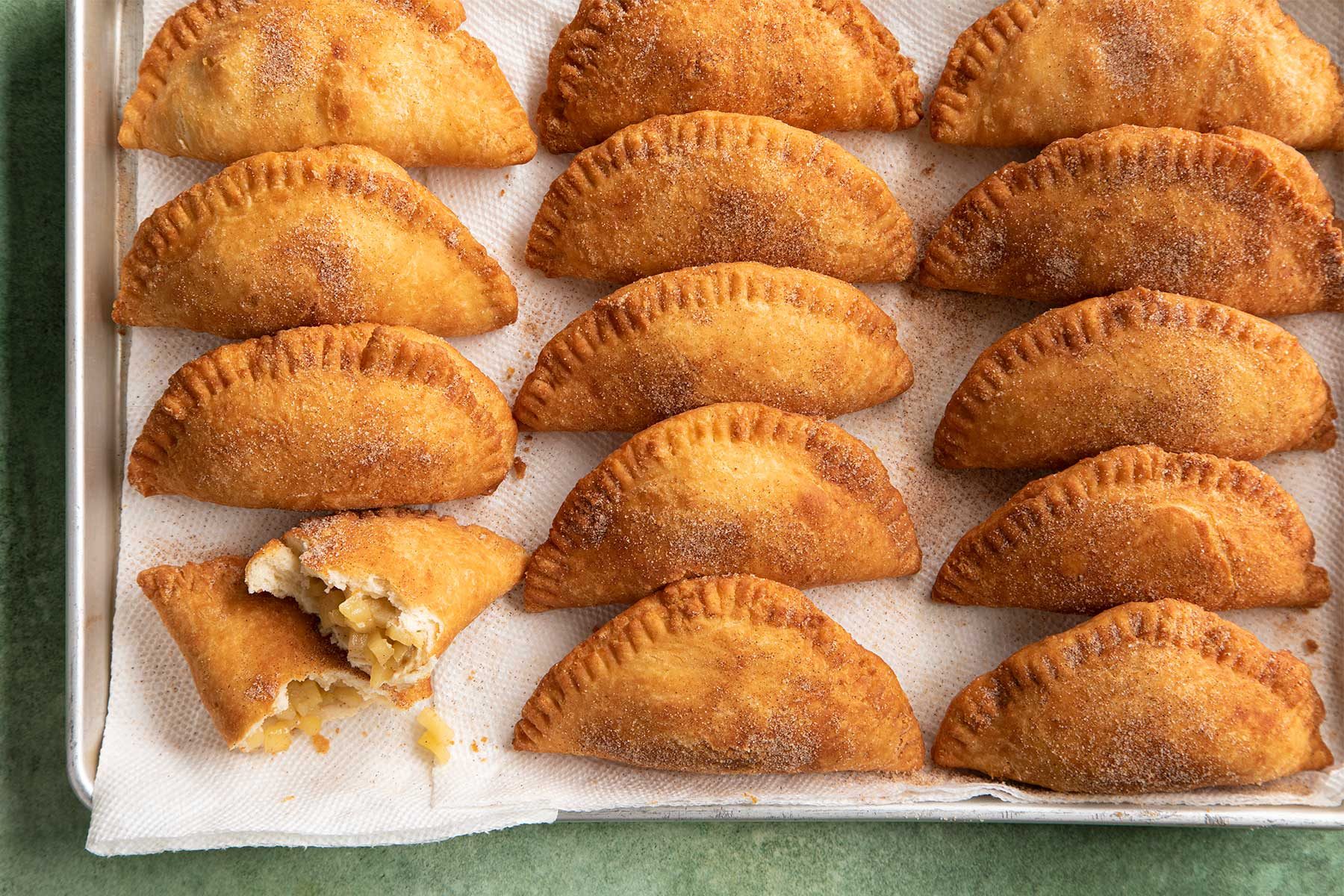 Overhead shot of eggs and milk being poured in a white bowl kept on top of a green marble countertop.Overhead shot of prepared fried apple pies kept on a black baking sheet with parchment paper on a green marble countertop.