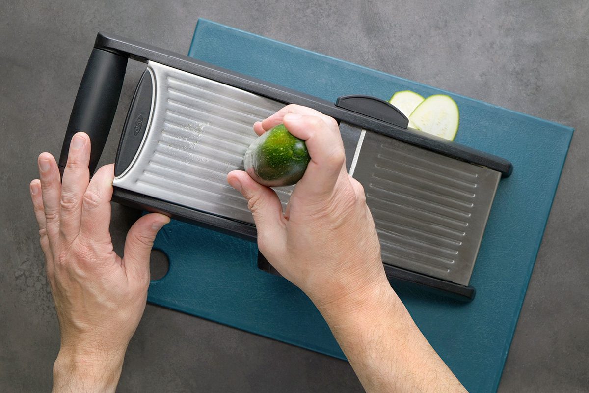 A person uses a mandoline slicer to slice a cucumber on a blue cutting board, with a few slices visible at the end of the slicer.