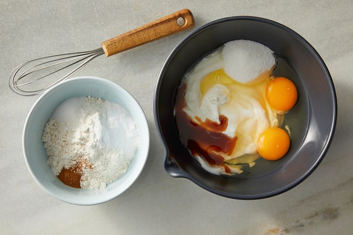 A whisk, a bowl of flour, cinnamon, baking powder, and a larger bowl with eggs, sugar, yogurt, and vanilla extract on a marble countertop, ready for baking.