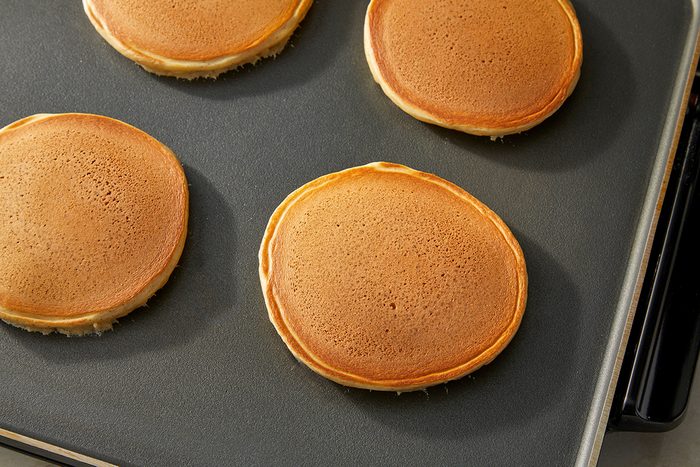 Four golden brown pancakes are cooking on a flat, nonstick griddle. The pancakes are evenly spaced and have smooth, round edges with lightly browned tops.