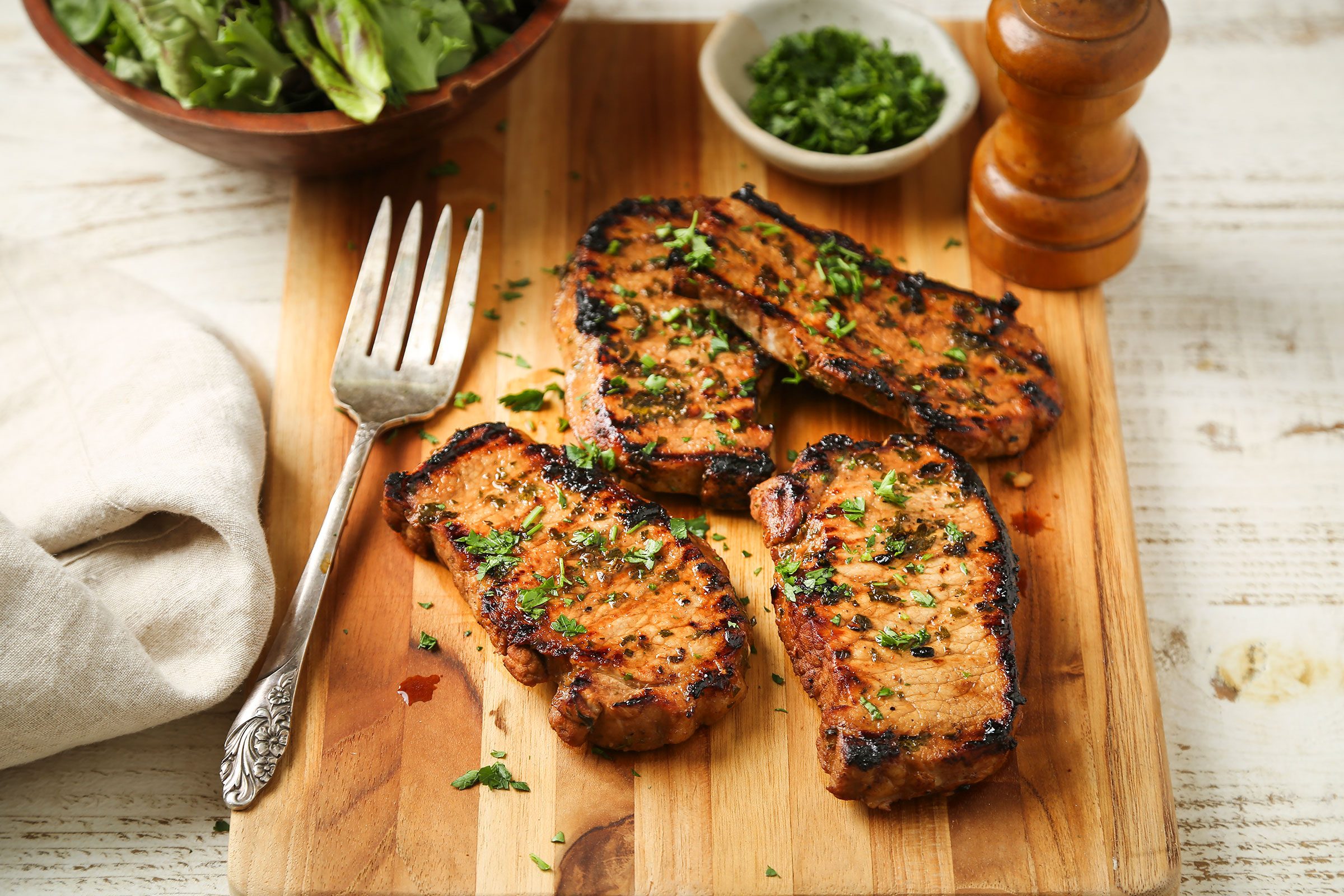 Grilled Boneless Pork Chops on a wooden board with a fork and some greens on side.