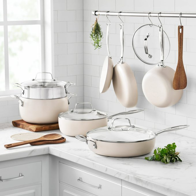 A set of cream-colored pots and pans with glass lids arranged on a white marble kitchen counter, with some cookware and lids hanging from a metal rack above. Fresh herbs and wooden utensils are nearby.
