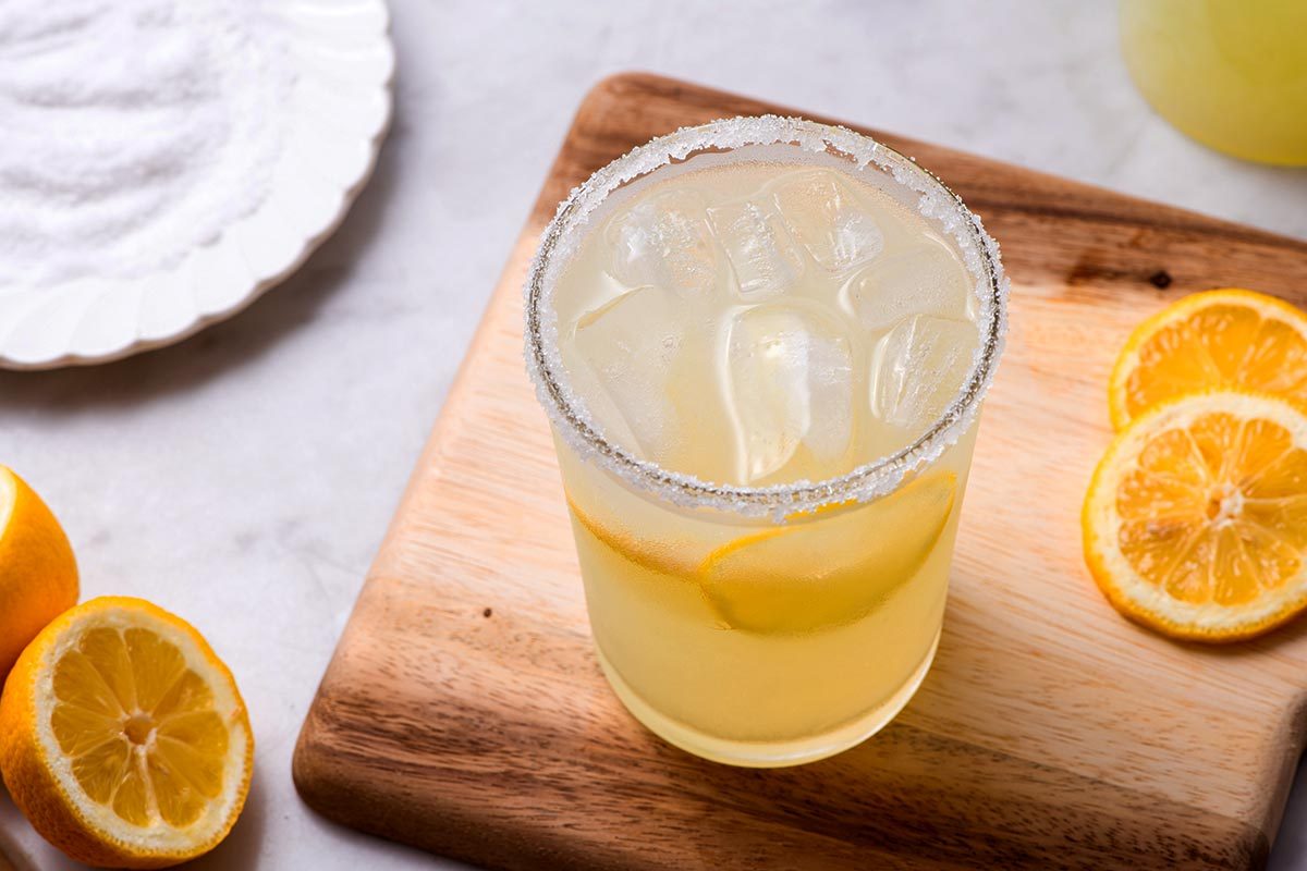 High angle shot of a glass of light yellow iced drink with lemon wedges on top of a cutting board, next to a plate of salt
