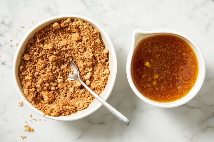 A bowl of crushed graham crackers with a spoon next to a small pitcher containing a melted butter and sugar mixture, both placed on a marble surface.
