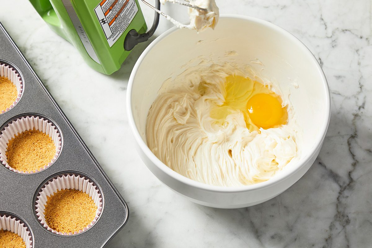 3/4 angle view shot of another small bowl; Beat cream cheese; sugar and vanilla until smooth; add egg; beat on low speed just until combined; A muffin tin lined with pink paper cups, filled with crumb mixture, is visible; A green electric mixer rests nearby; all set on a marble surface
