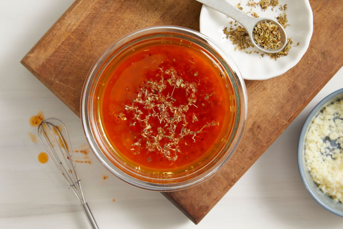 Overhead shot of Sub Oil and Vinegar Drizzle; In a small glass bowl placed over wooden board; whisk together all of the ingredients in bowl; a spoon over a small plate; all set on a cream surface;