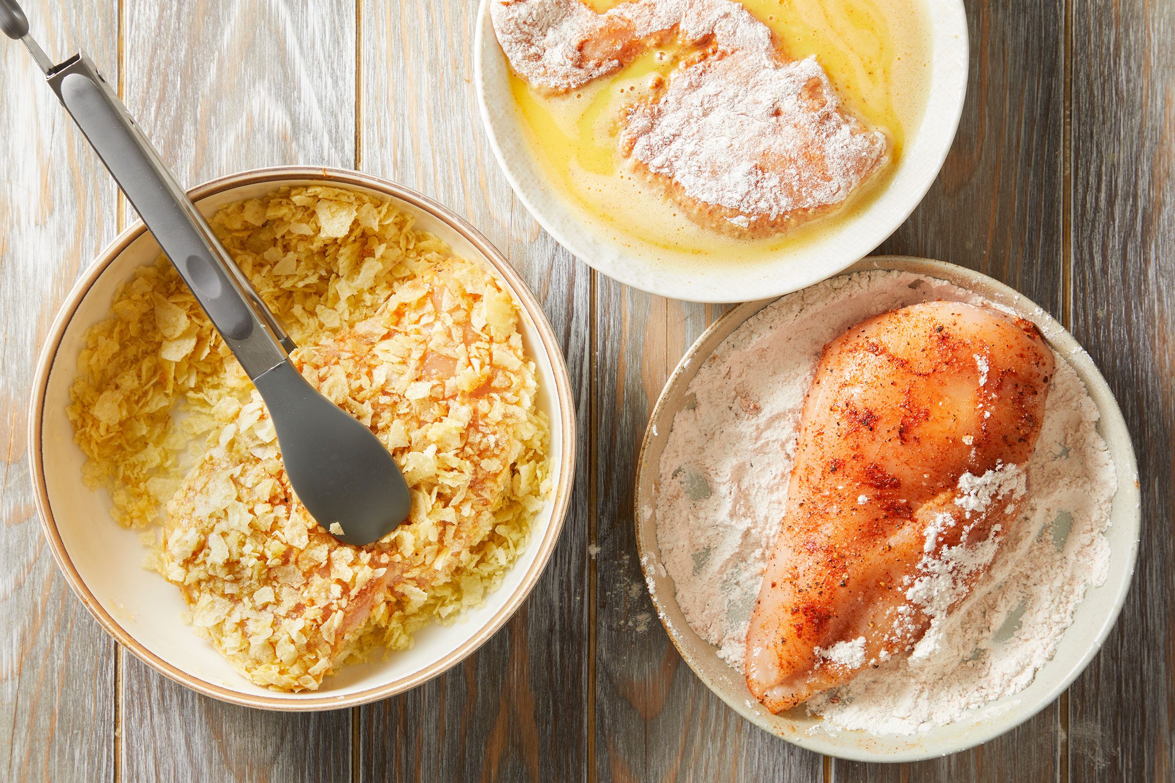 Chicken breasts coated in the flour mixture with some dipped into the beaten egg with a bowl of crushed potato chips