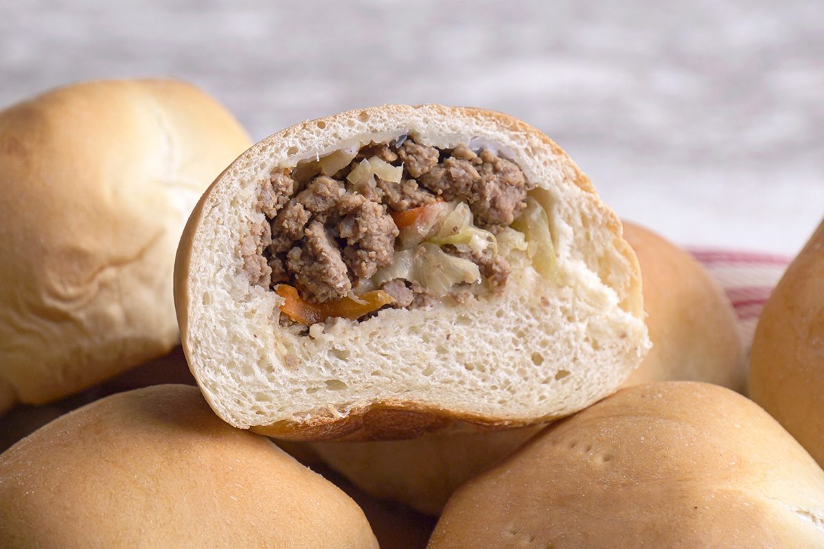 A close-up of a baked bun sliced open, revealing a filling of ground meat and vegetables. Several whole buns are stacked around it on a light surface.