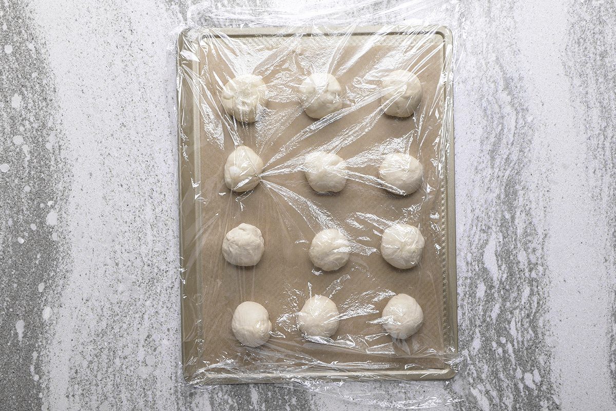 A baking sheet with twelve dough balls covered in plastic wrap, set on a marbled countertop.