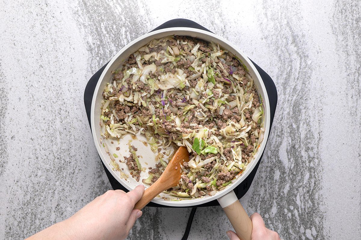 A person stirs a pan filled with a mixture of ground meat, chopped cabbage, and vegetables on a stovetop.