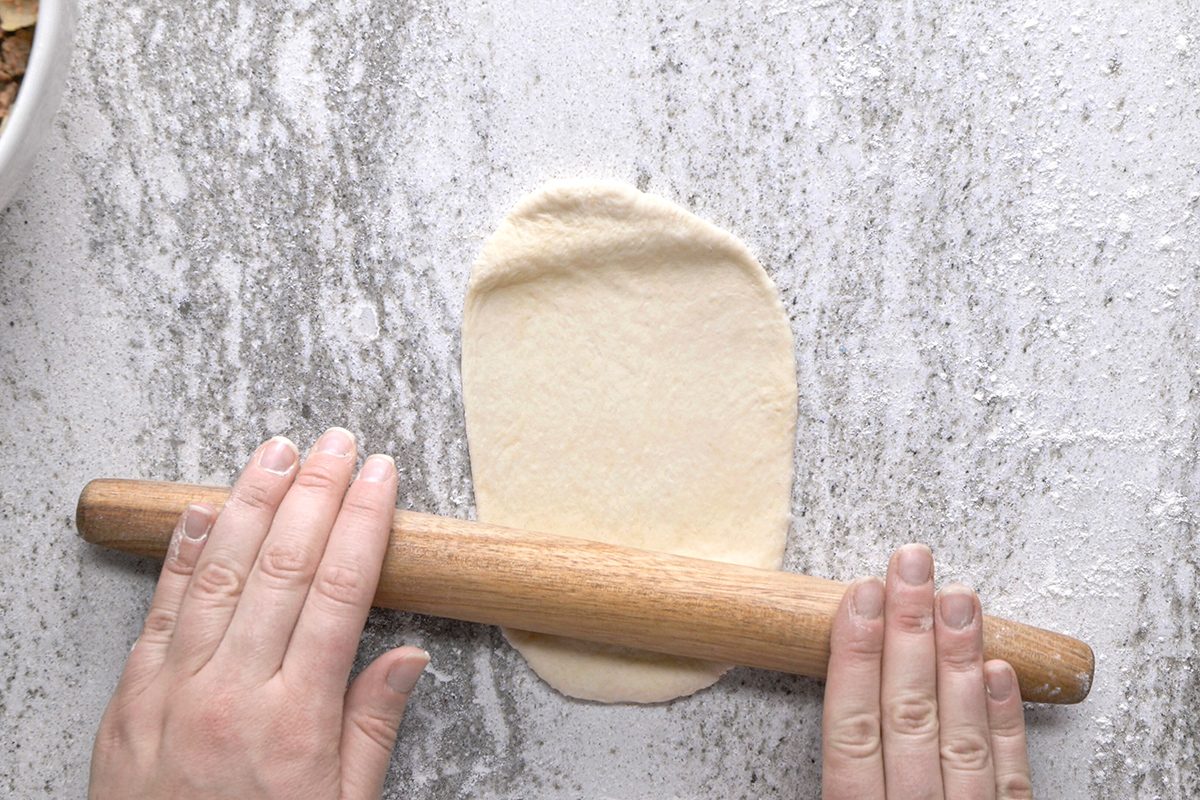 A person rolls out a small oval piece of dough with a wooden rolling pin on a floured, gray countertop.