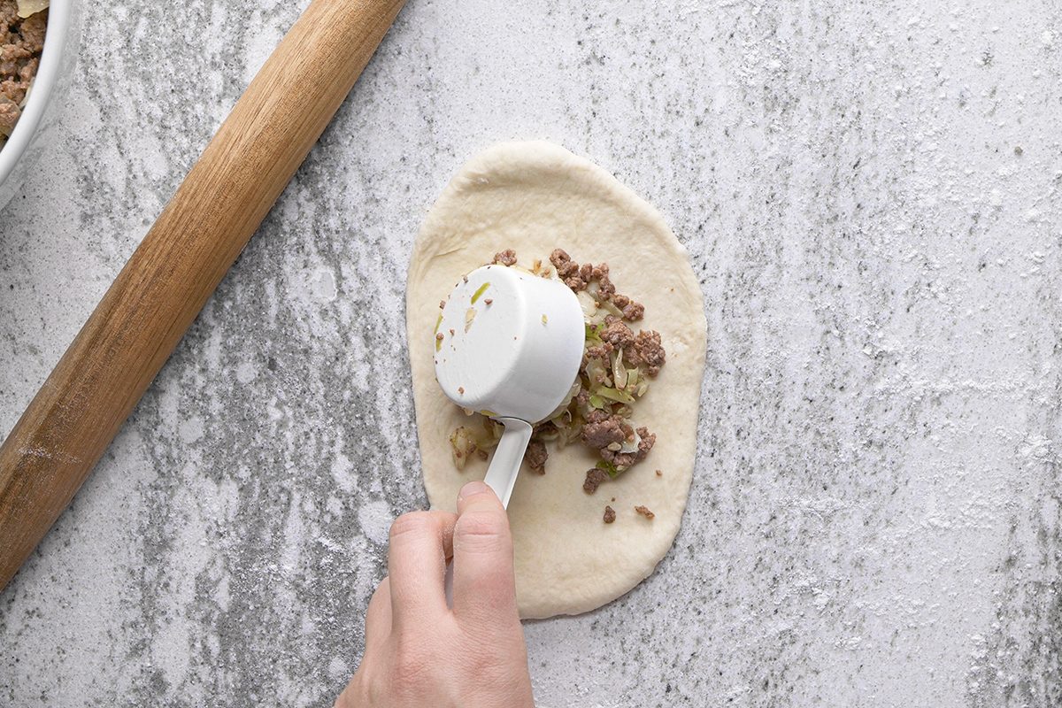 A hand holds a measuring cup, scooping a ground meat and vegetable mixture onto an oval-shaped piece of dough on a floured surface, with a rolling pin nearby.