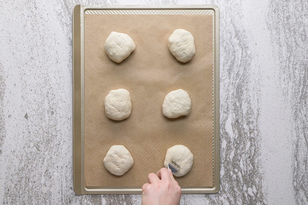 A hand presses down on one of six round dough balls arranged on a parchment-lined baking sheet, ready for baking.