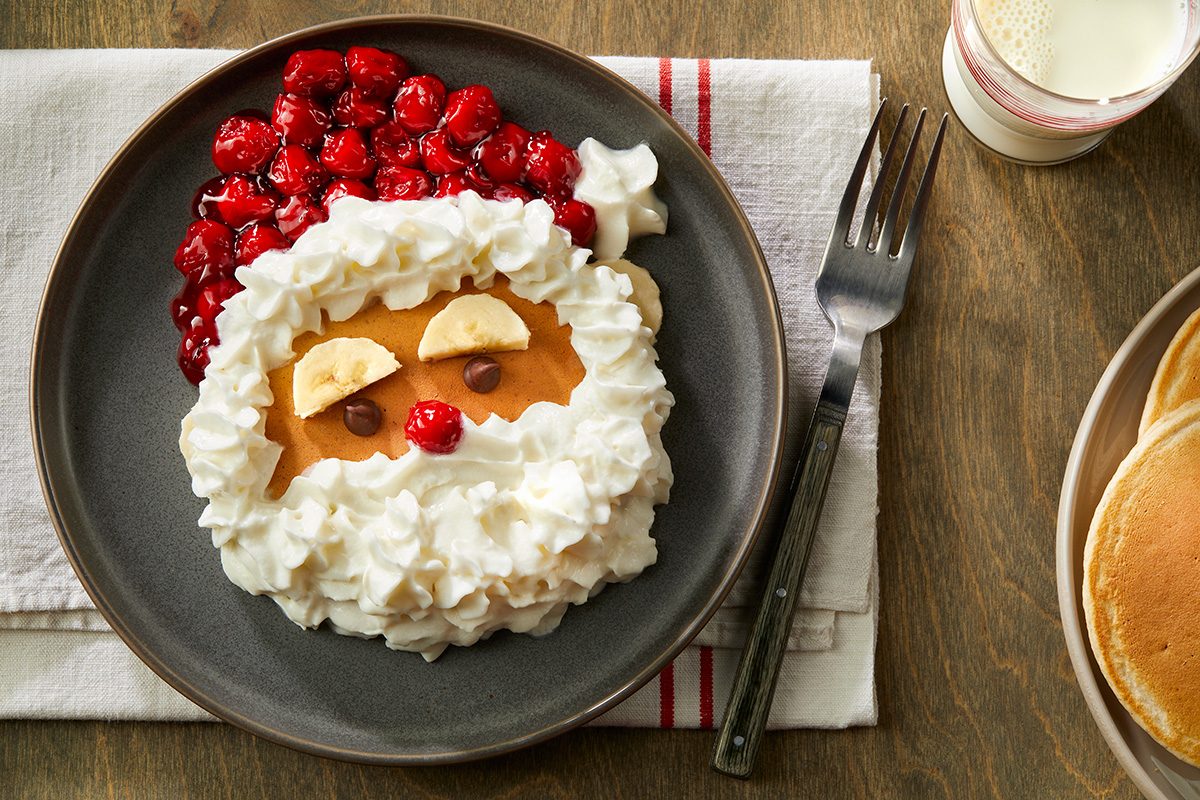 Overhead shot of Santa pancakes; served on a black ceramic plate; with a glass of milk and a fork on a napkin