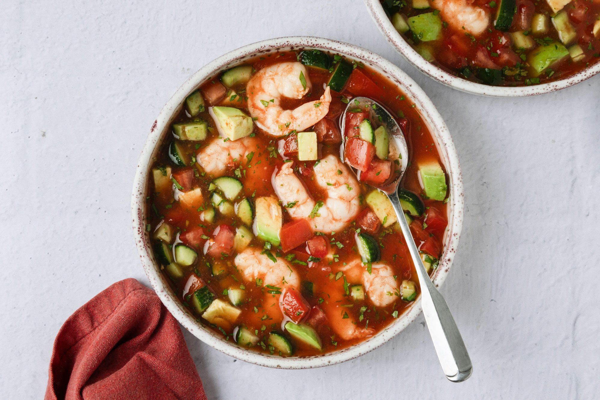 overhead shot of a bowl filled with Shrimp Gazpacho, a silver spoon rests against the side of the bowl, in the background, another bowl is partially visible, a napkin is placed near the main bowl