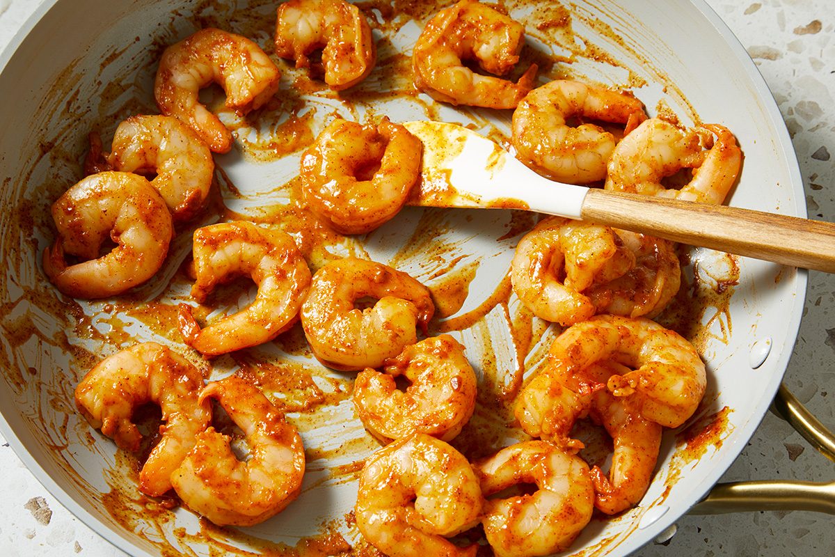 Shrimp coated in a reddish sauce cooking in a white skillet, being stirred with a wooden spatula.