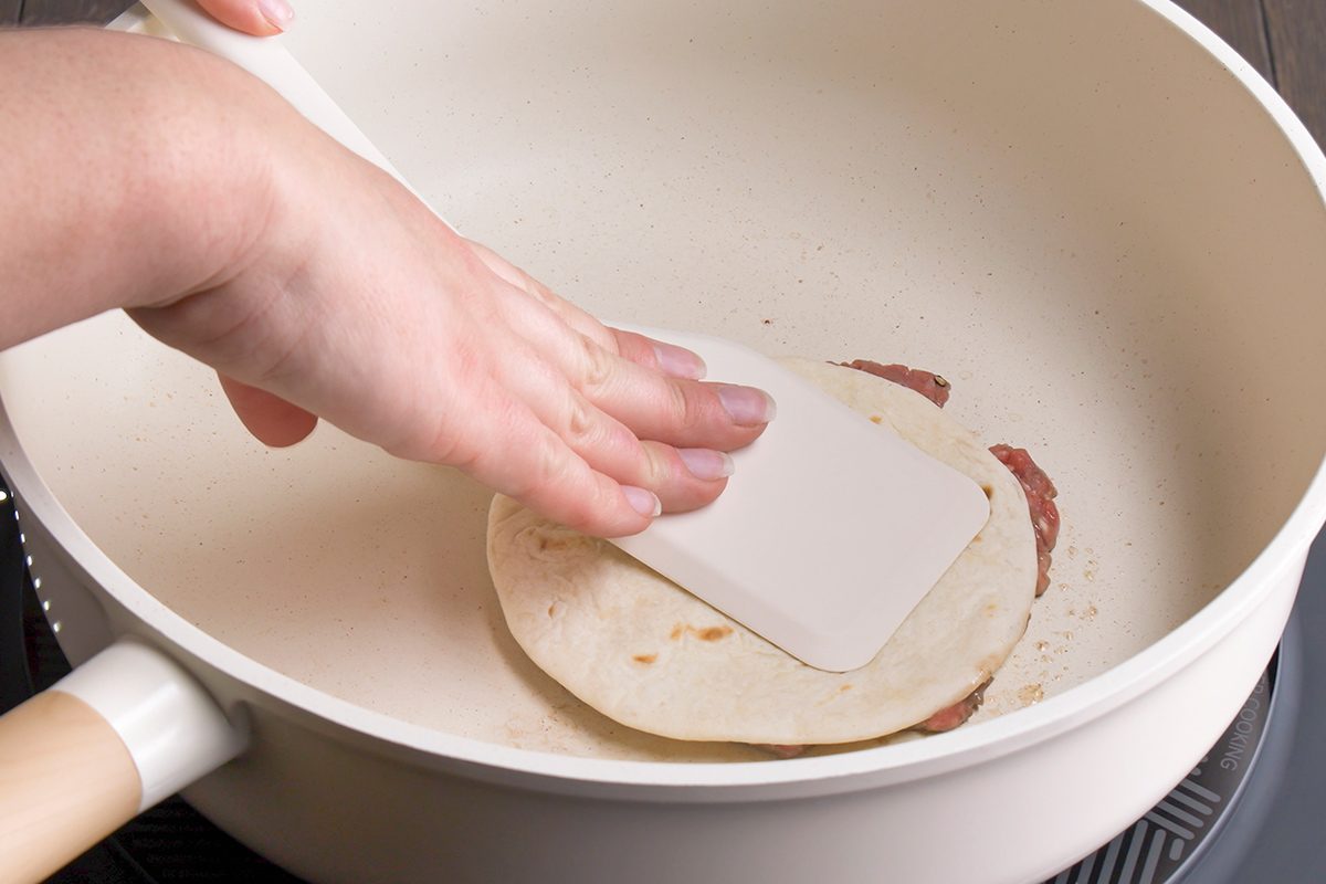 A hand presses a white spatula onto a tortilla in a white frying pan on a stovetop, cooking the tortilla.