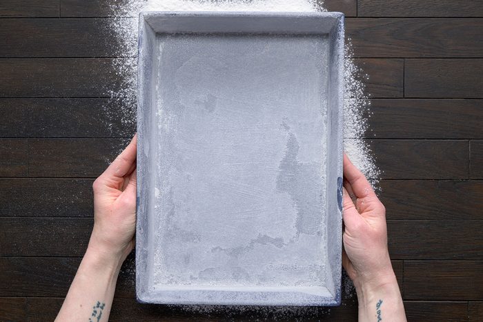overhead shot of two hands are shown holding a rectangular baking pan over a dark brown wooden surface; dusting flour