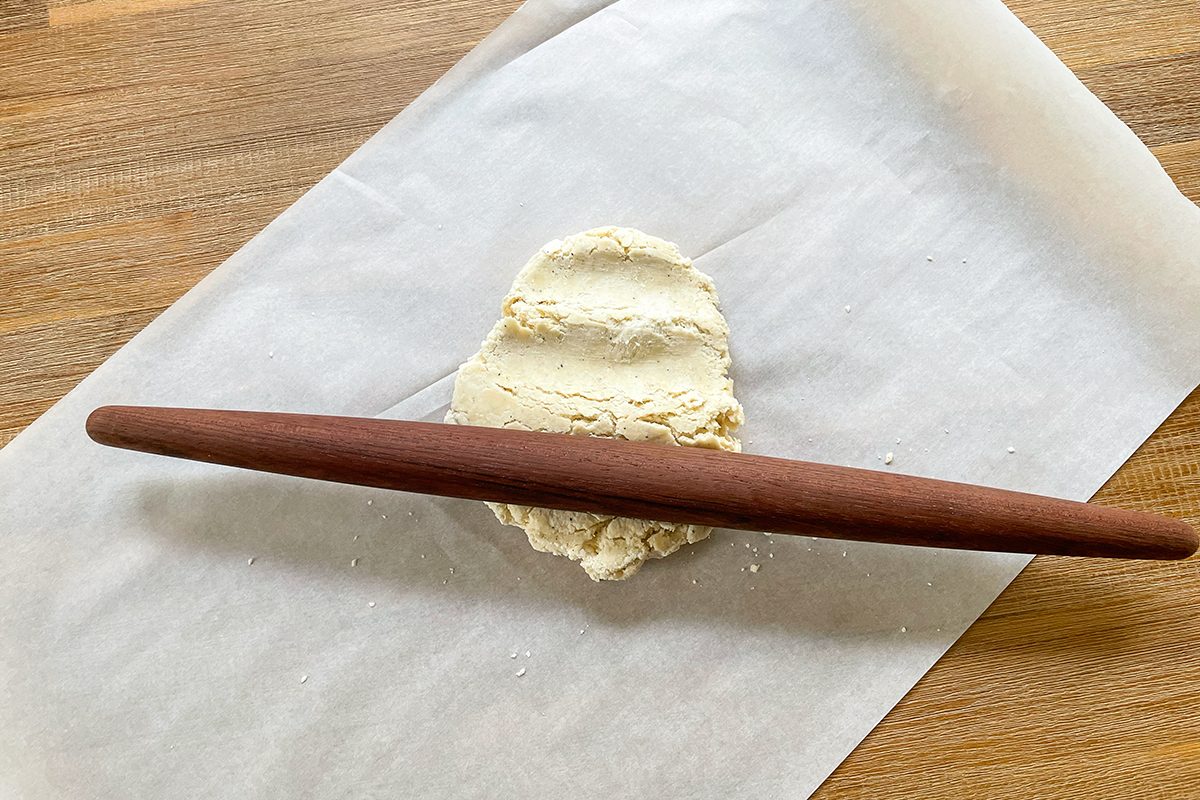 A rolling pin is flattening a piece of dough on parchment paper, placed on a wooden surface.