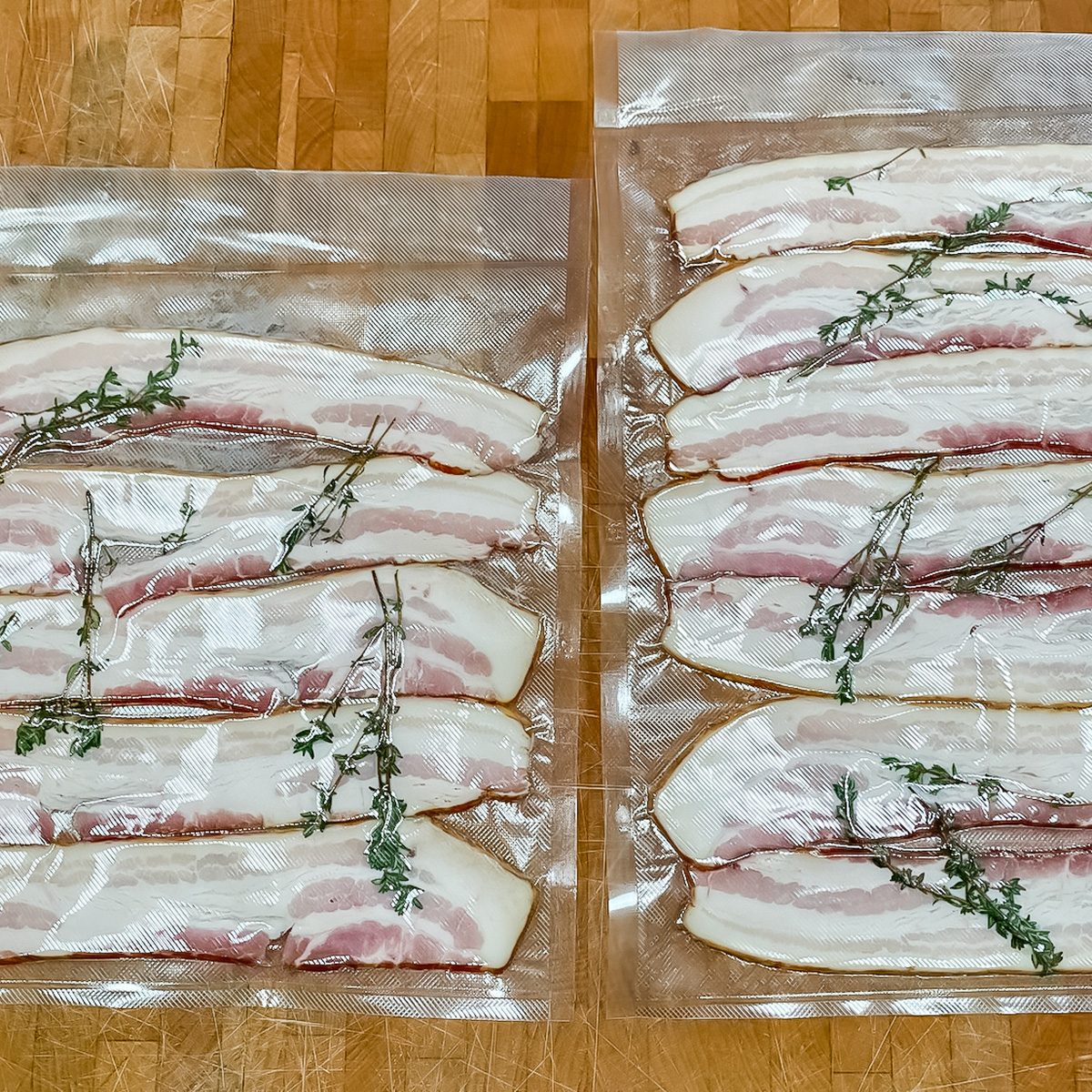 Two vacuum-sealed bags each contain several strips of raw bacon with sprigs of fresh herbs, arranged on a wooden surface.
