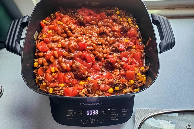 A digital cooker with a timer reading 38 minutes shows a dish of mixed beans, corn, rice, and diced tomatoes in a tomato-based sauce. The meal is colorful and appears to be a hearty stew or chili.