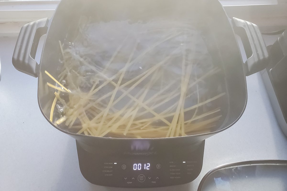 A pot of boiling water on a stovetop with uncooked spaghetti partially submerged, steam rising above. The control panel on the pot displays the number 12.