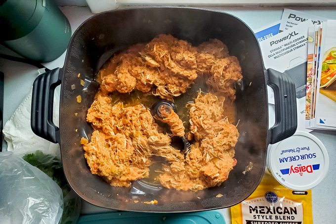 Shredded chicken cooking in a black skillet, surrounded by various ingredients and packaging, including sour cream, Mexican style cheese, and leafy greens. Steam is visible rising from the chicken.
