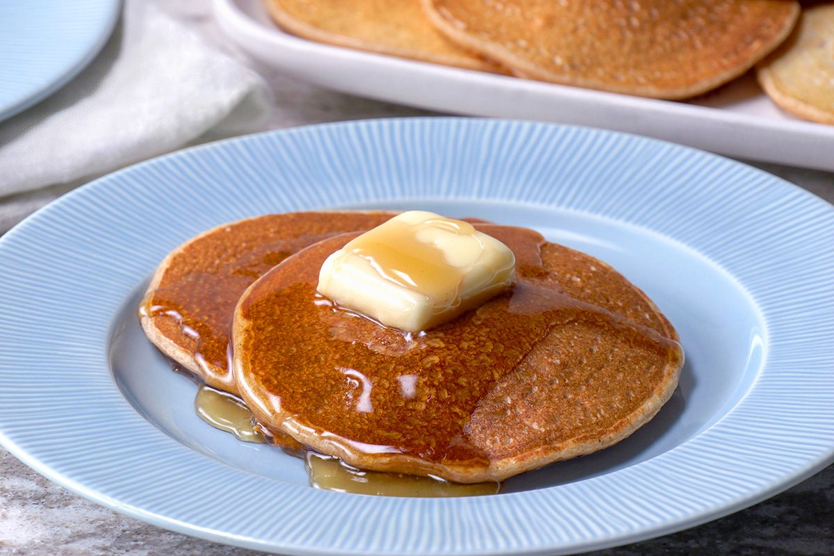 Two pancakes on a blue plate, topped with a pat of butter and drizzled with syrup. More pancakes are visible on a platter in the background.