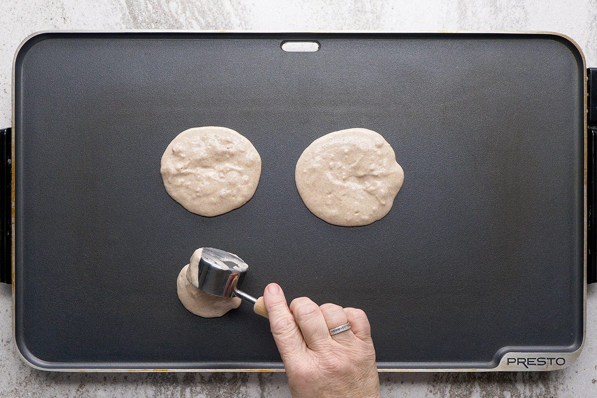 A hand pours pancake batter from a metal measuring cup onto a black griddle, forming a third pancake next to two already cooking.