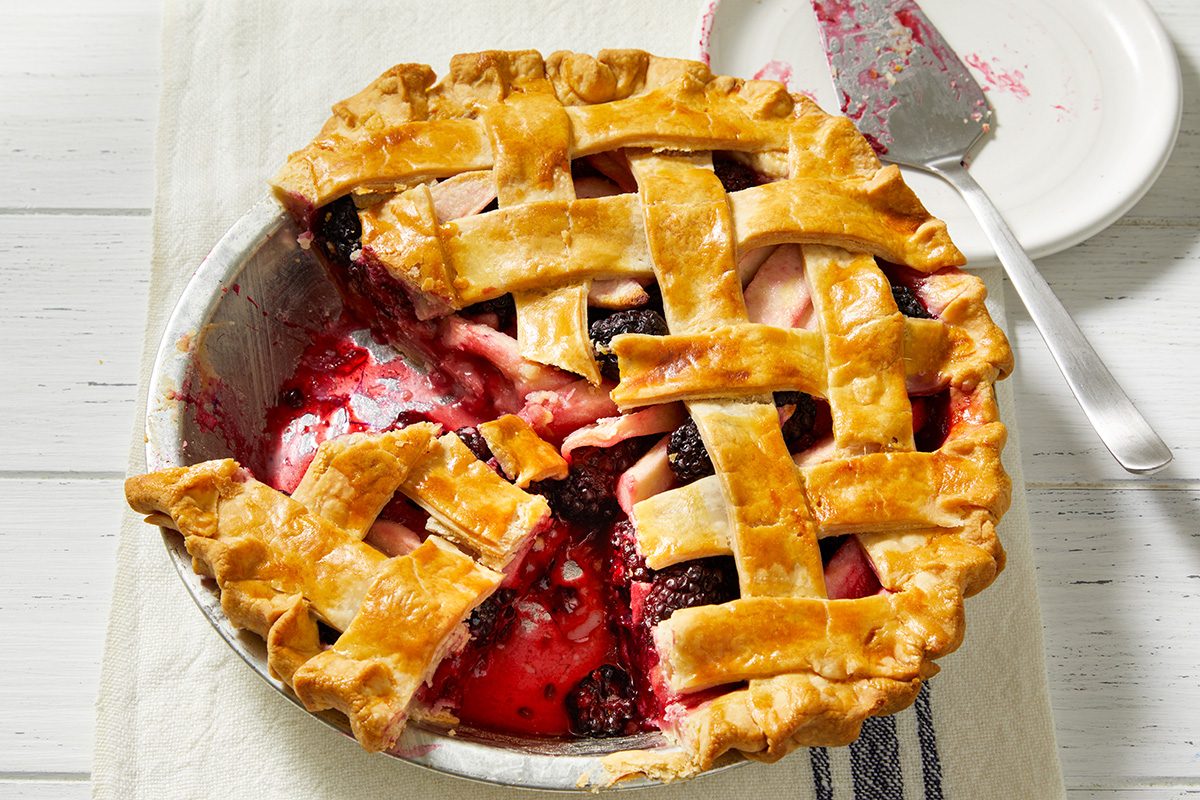 A baked lattice-top pie with a slice missing, showing a filling of berries and fruit. The pie sits on a white surface with a pie server and plate nearby, some filling spilled onto the dish.