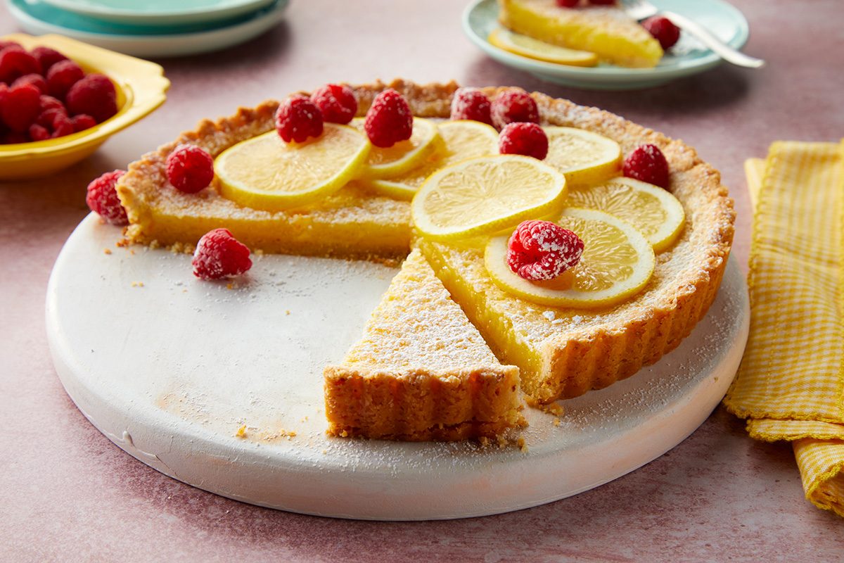 A lemon tart with a slice missing, topped with lemon slices and raspberries, sits on a white round serving board. A yellow napkin and plates with more tart and raspberries are in the background.