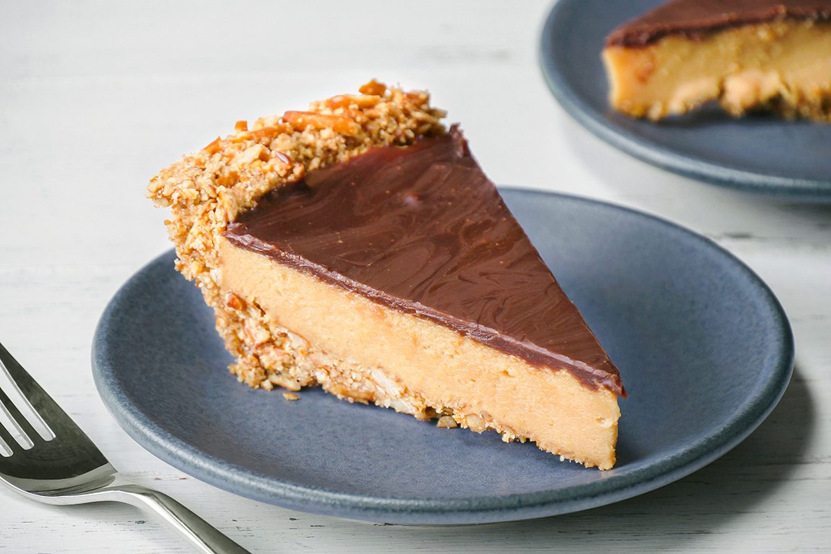 A slice of peanut butter pie with chocolate topping and a crumbly crust on a blue plate, with a fork beside the plate and another slice visible in the background.