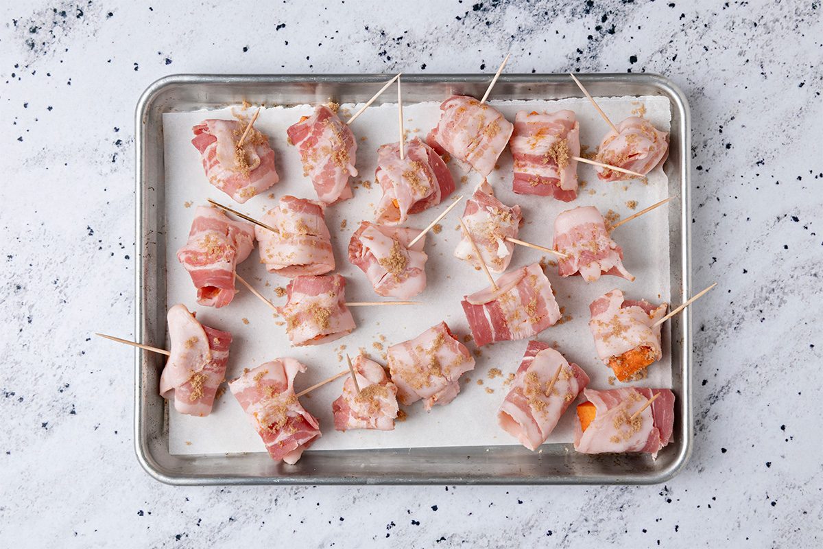 A metal baking tray lined with parchment paper holds several pieces of bacon-wrapped appetizers secured with toothpicks, sprinkled with brown sugar, ready to be baked.