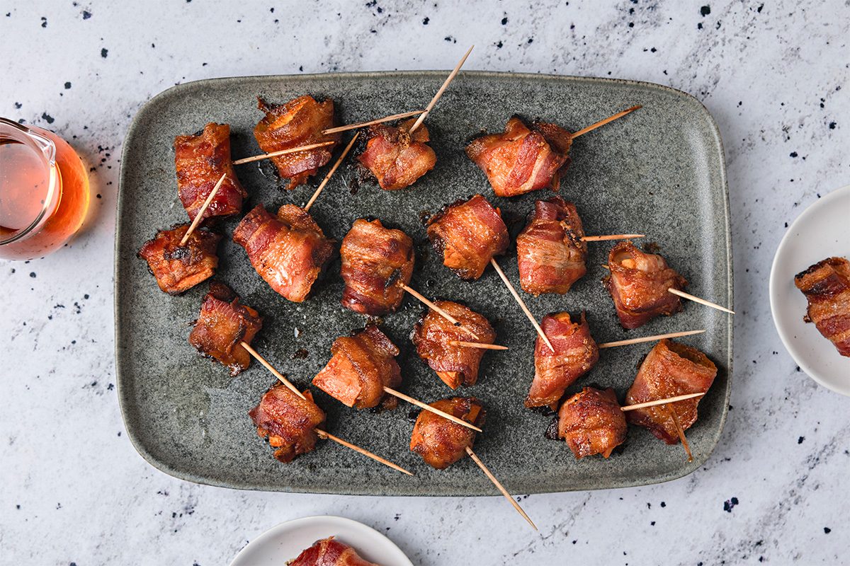 A grey plate holds multiple bacon-wrapped appetizers secured with toothpicks on a light, speckled surface. A glass of beverage and small white plates with more appetizers are nearby.