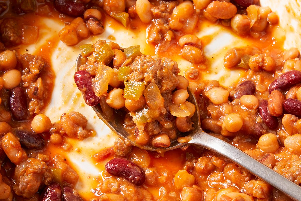 A close-up of a spoonful of chili with ground meat, red kidney beans, white beans, green peppers, and tomato sauce, surrounded by more chili in a dish.