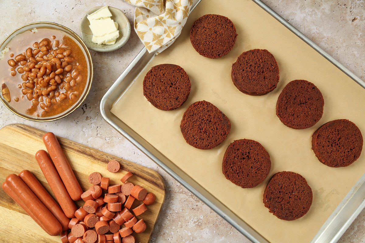 Overhead shot of kitchen top; Slice the brown bread into 8 portions and place them onto a greased baking sheet; bake according to the package directions; a bowl of baked beans and a wooden cutting board with the chopped hot dog are nearby with a napkin; all arranged on a marble surface;