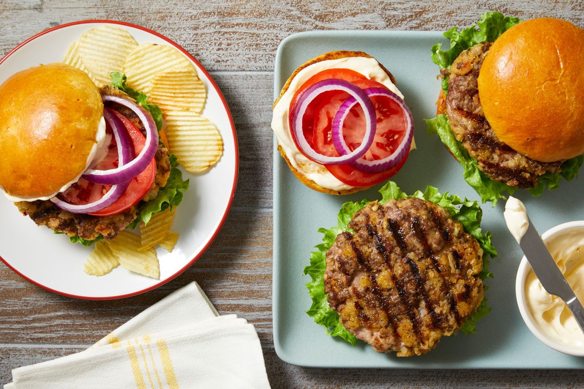 overhead shot of Beef and Pork Burger displayed in the foreground on a white plate with a red rim; in the background, additional burgers can be seen resting on a rectangular plate, the setting features a rustic wooden table