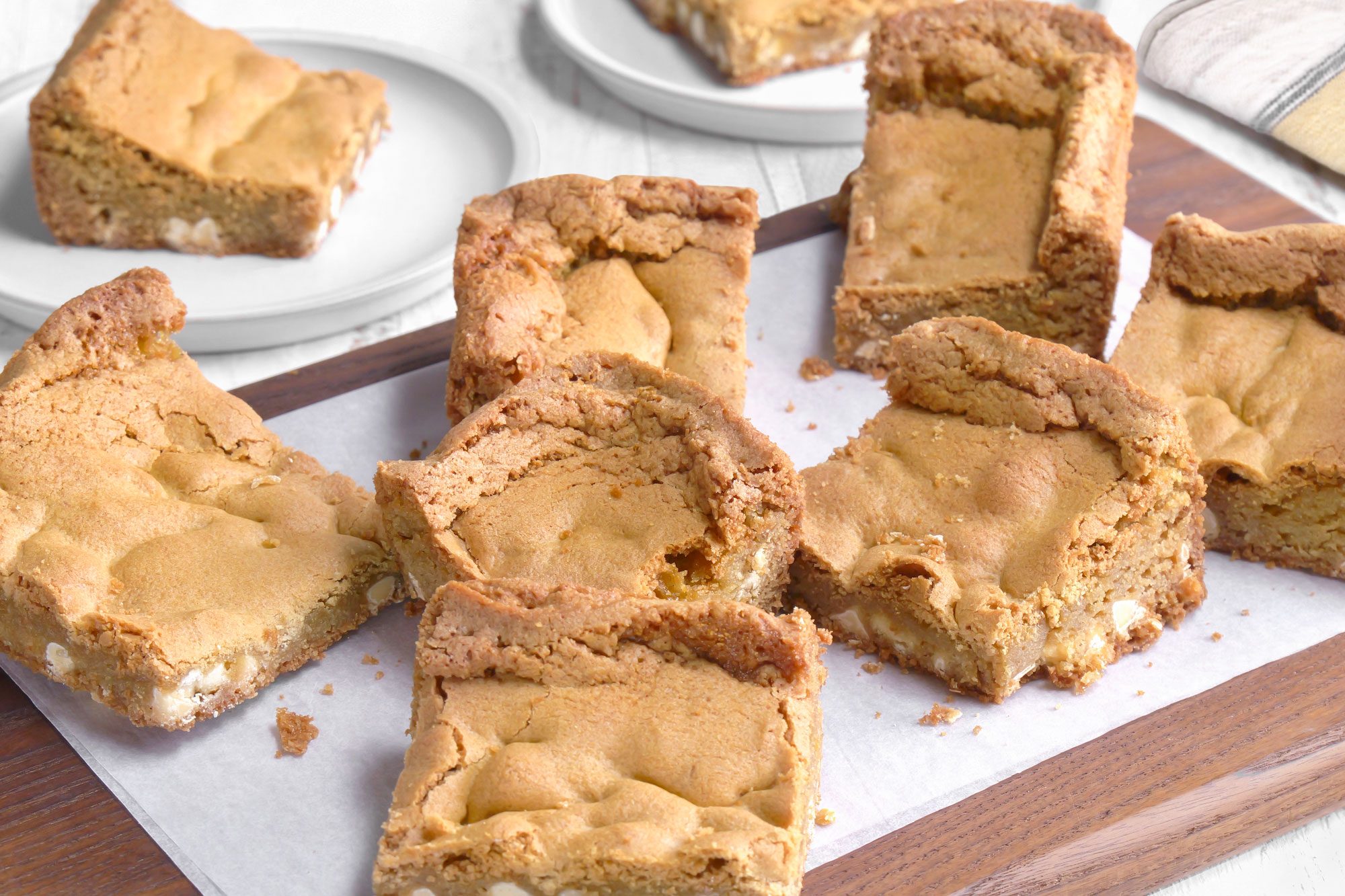 3/4th shot of Squares of golden brown blondies are arranged on a parchment lined wooden cutting board with a few pieces on nearby white plates, all set on a white table