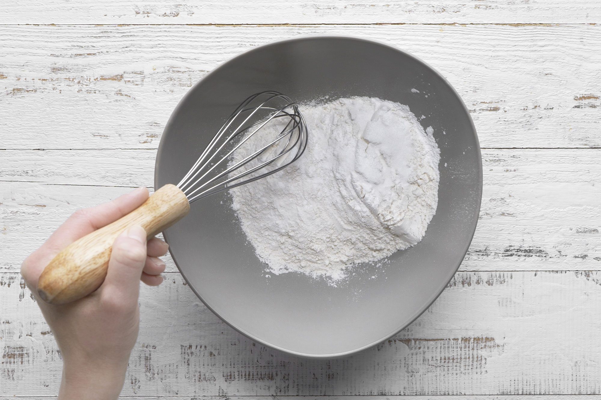 overhead shot of a hand holding a whisk mixes flour in a gray bowl on a white wooden surface
