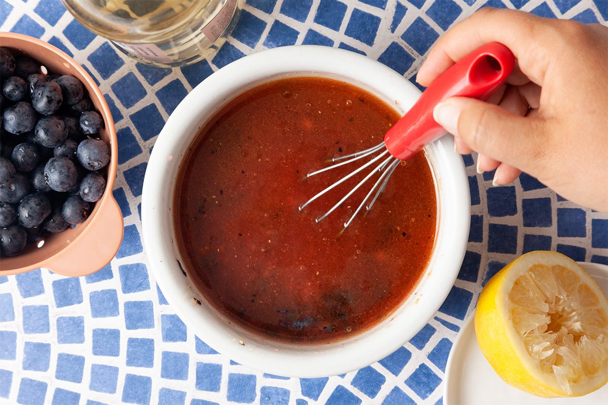 A hand uses a small whisk to mix a bowl of reddish-brown sauce. Nearby are a cup of blueberries, a glass of liquid, and a halved lemon on a blue and white tiled surface.