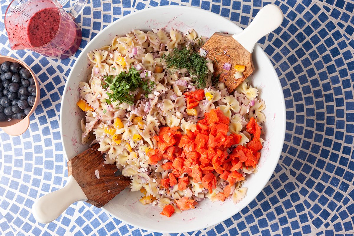 A large white bowl filled with pasta salad, chopped vegetables, herbs, and orange smoked salmon sits on a blue mosaic table. Wooden utensils, a drink, and a small bowl of blueberries are nearby.