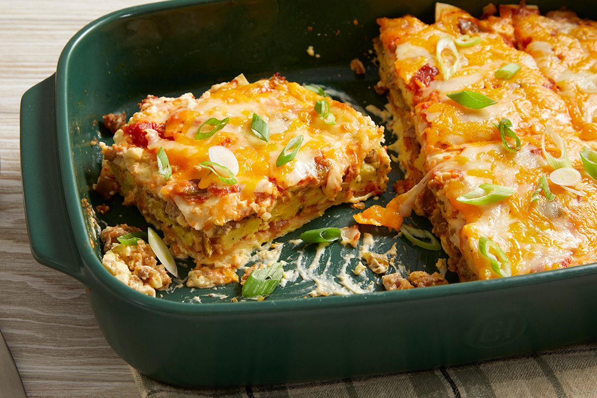 A green baking dish containing cheesy, golden-brown lasagna with a large square piece cut out and lifted slightly. The lasagna is topped with chopped green onions.