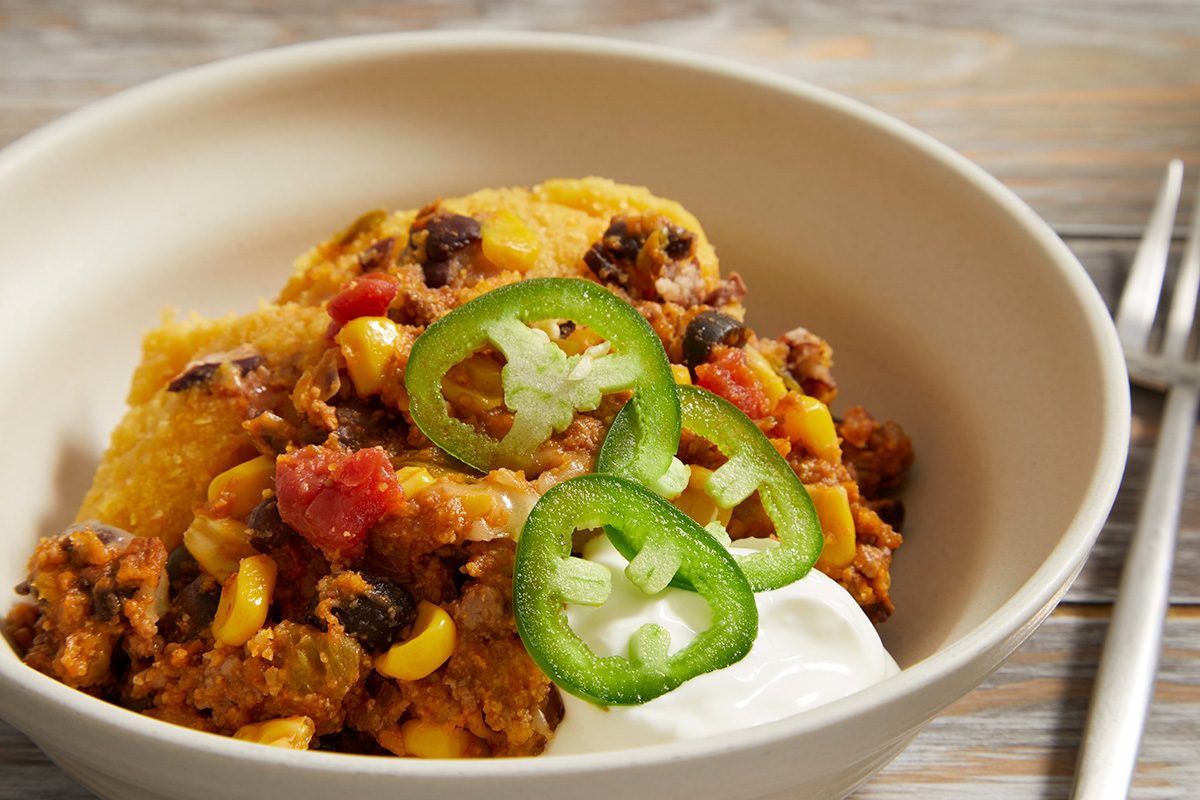 A bowl of chili topped with cornbread, sliced jalapeños, and a dollop of sour cream, served on a wooden table with a fork nearby.