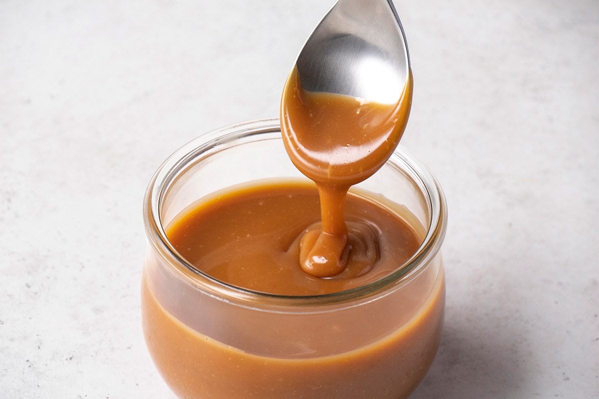 A spoon drizzles thick, golden caramel sauce into a small glass jar filled with caramel, set on a light-colored surface.