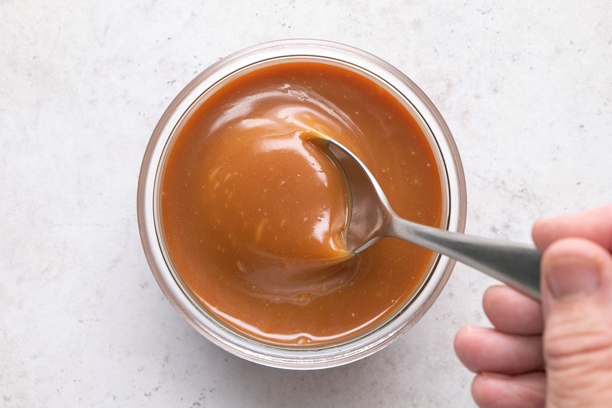 A hand holds a spoon scooping thick, smooth caramel sauce from a small glass bowl on a light-colored surface.