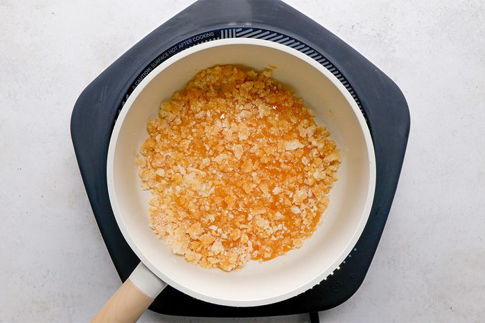 A saucepan with light brown, crumbly melted sugar sits on a black hot plate against a light background. The sugar is partially liquefied and has a grainy texture.