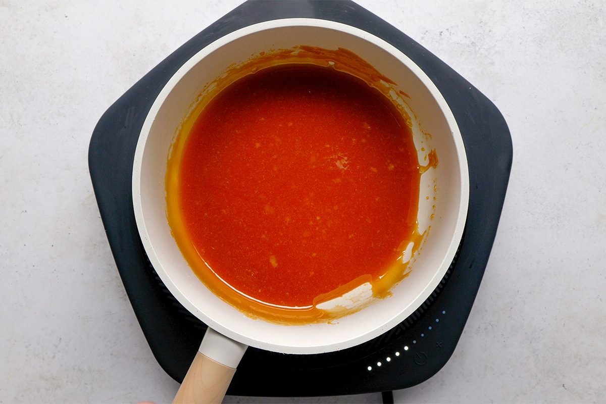 A white saucepan with an orange-red sauce sits on a black induction cooktop against a light gray background. The sauce appears smooth and glossy, coating the bottom and sides of the pan.