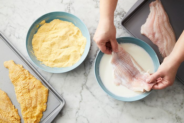 Hands dip a raw fish fillet into a bowl of milk, next to another bowl filled with cornmeal. On a baking sheet, one fillet is breaded and another is unbreaded, all on a marble countertop.
